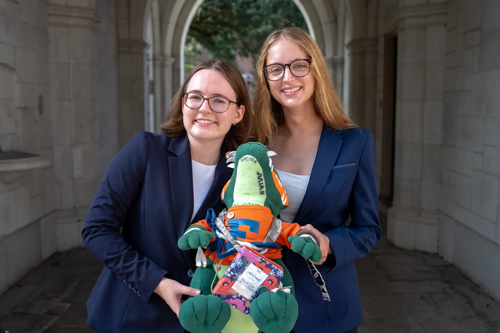 Two members in navy blazers hold a plush gator under a stone archway.