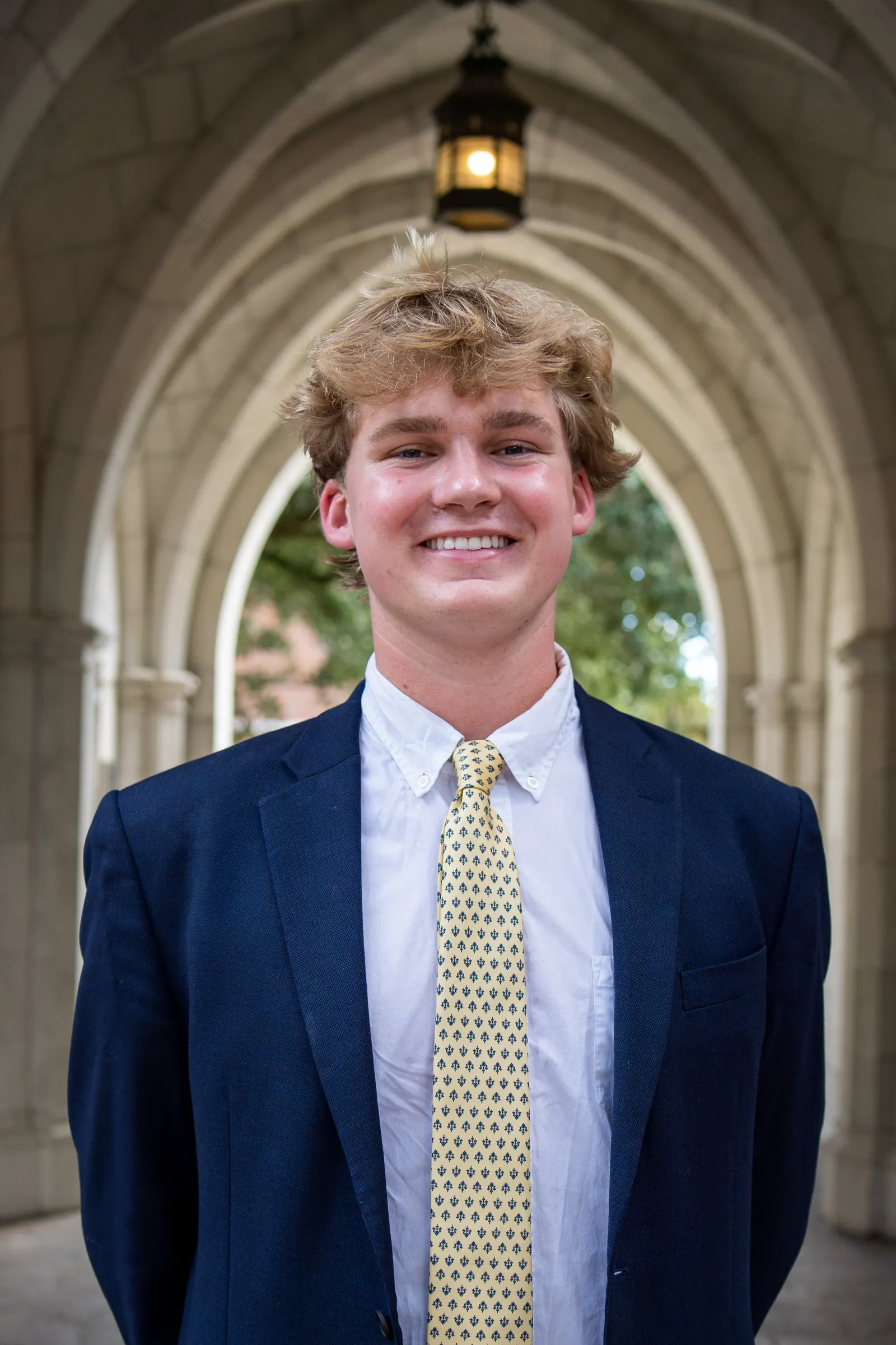 Headshot of a young man in a navy blazer and gold tie under a stone archway.