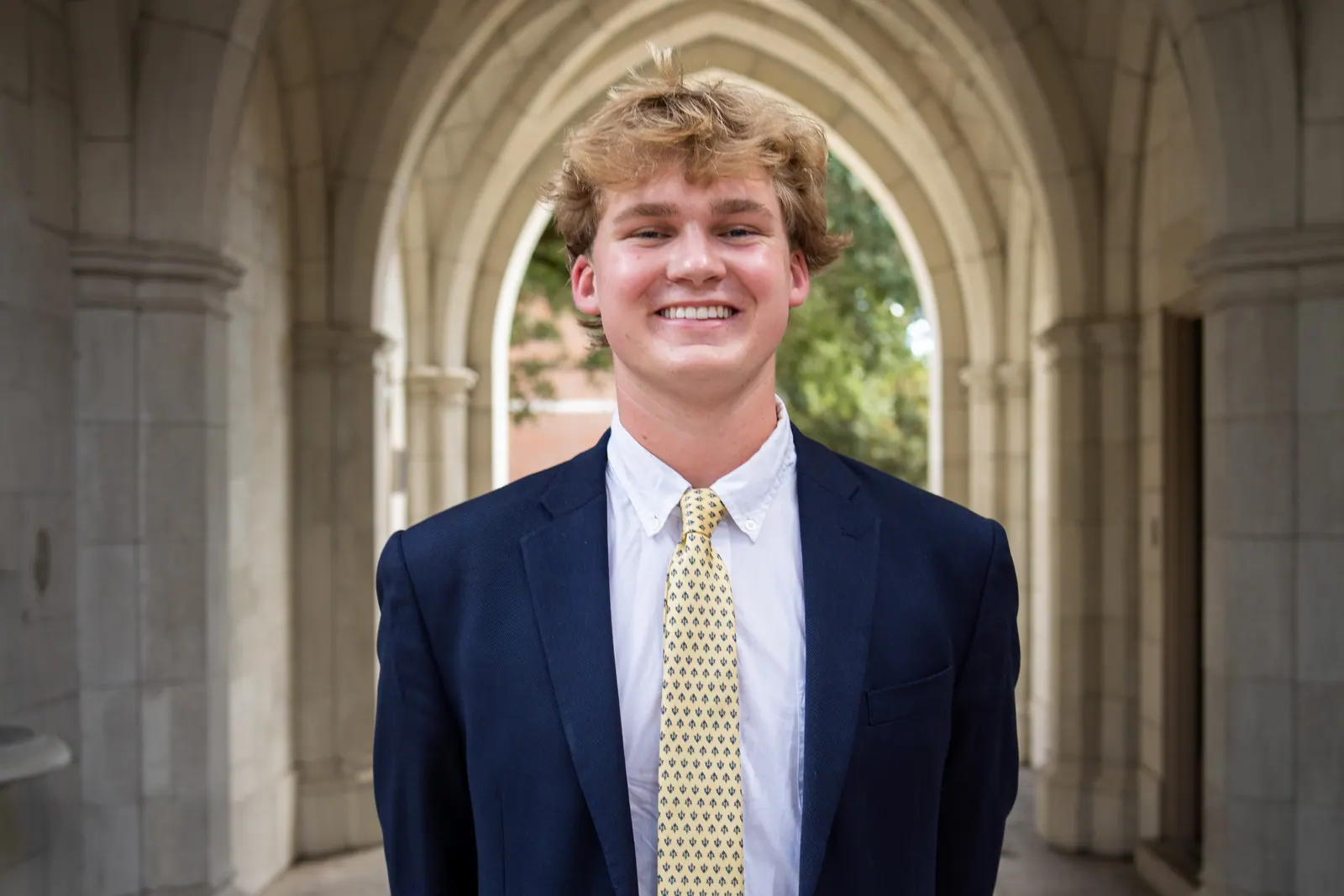 Wider headshot of a young man in a navy blazer and gold tie in a stone corridor.
