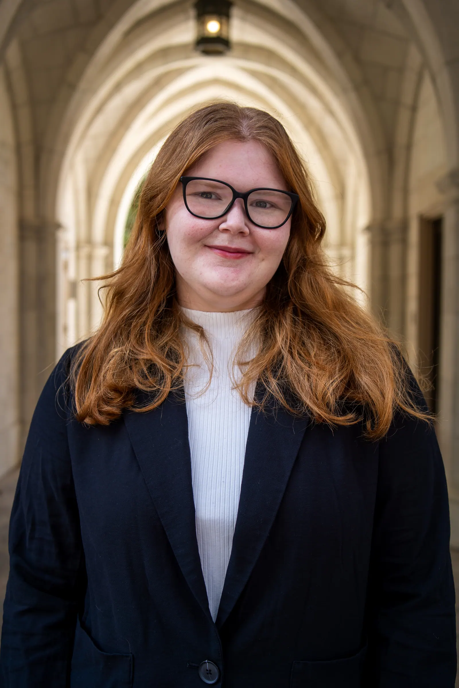 Headshot of a young woman with glasses in a black blazer under a stone archway.