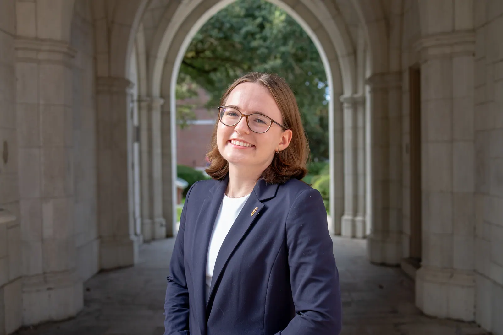 Wide headshot of a young woman with glasses in a navy blazer in a stone corridor.