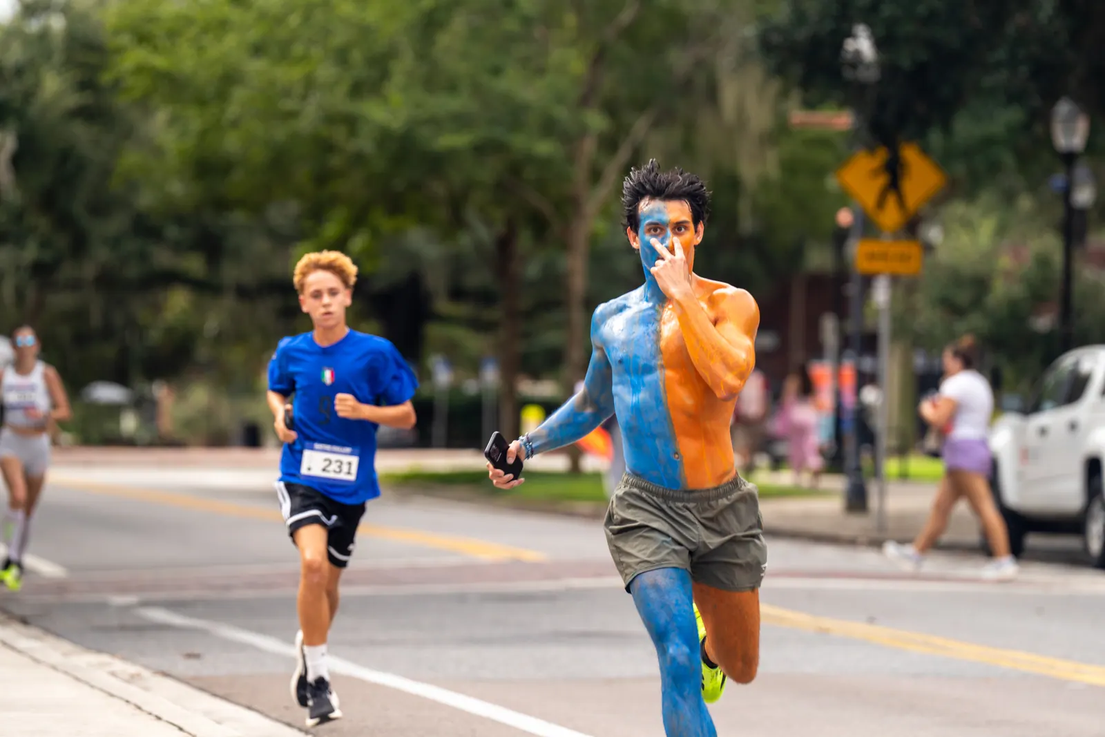 A runner painted half blue and half orange points ahead while sprinting down the road.