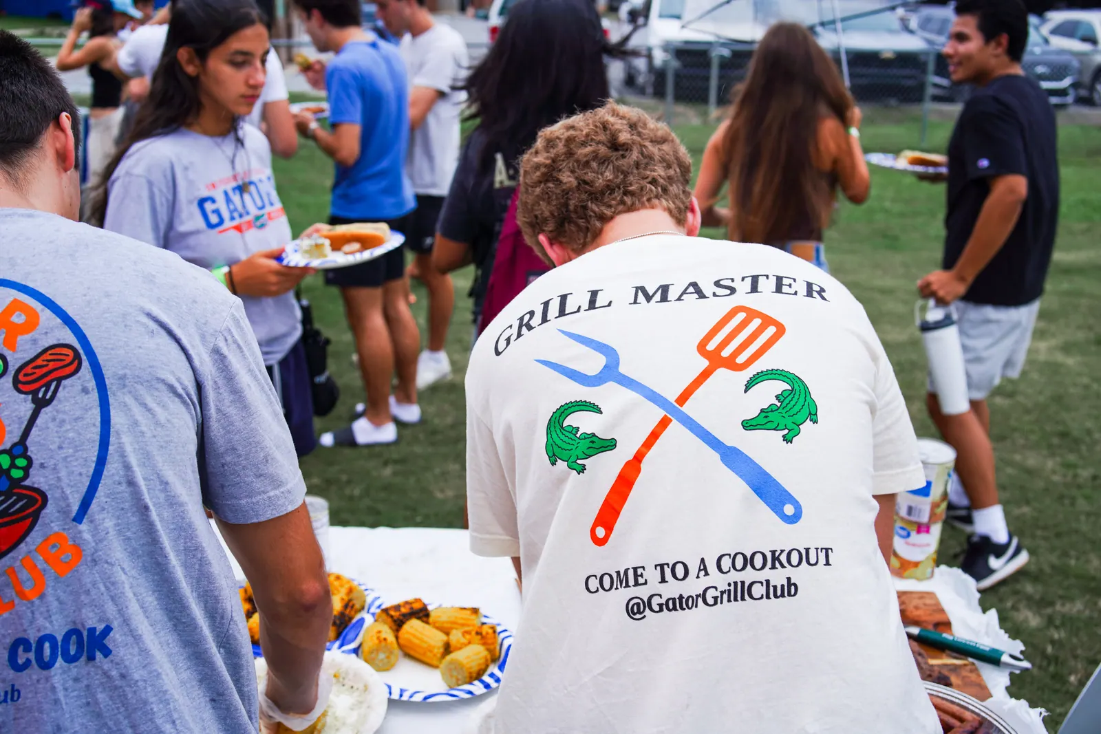 A student in a 'Grill Master' Gator Grill Club shirt serves food as the line moves along the table.
