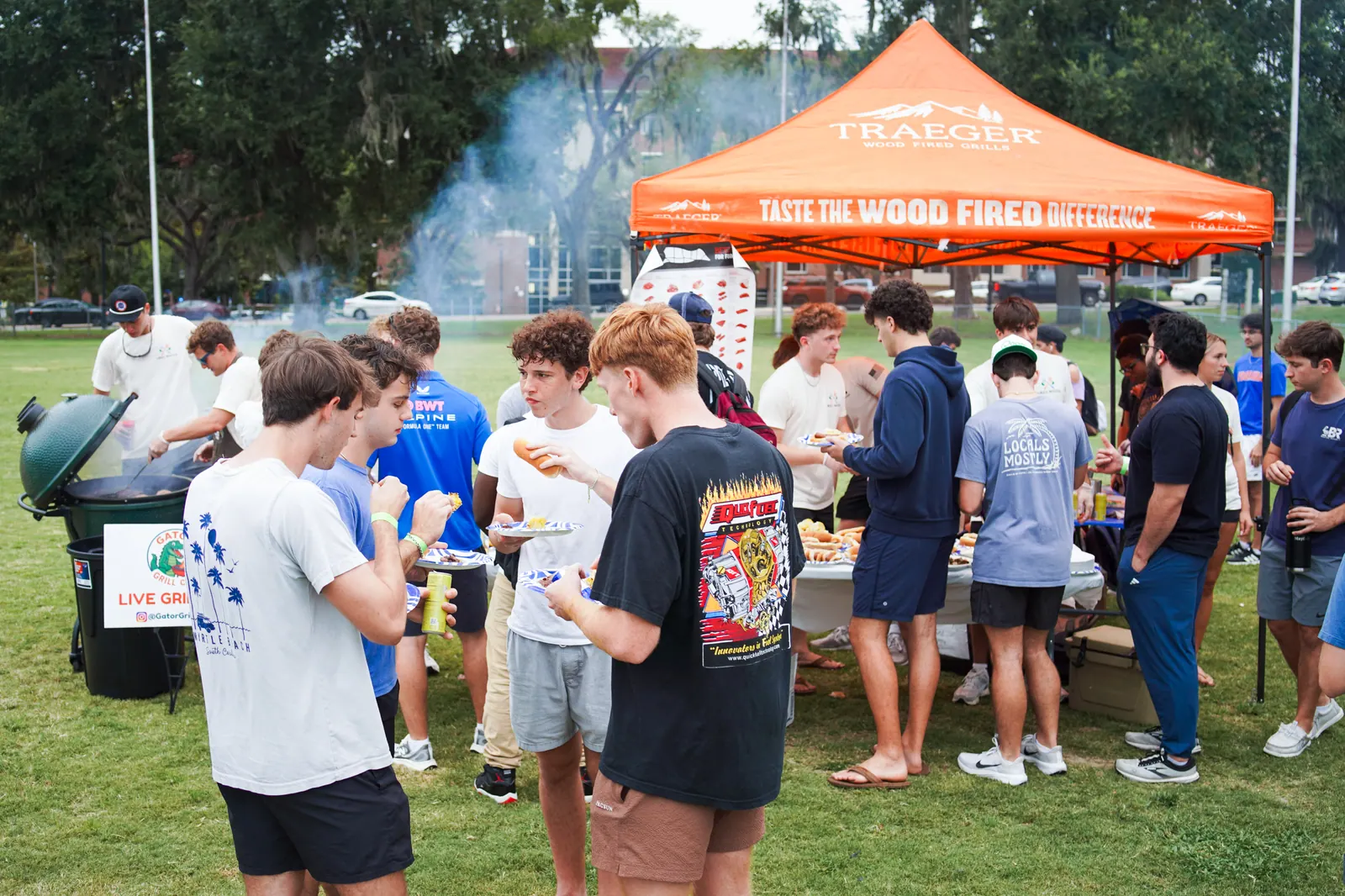 Students crowd around the serving tables beside the orange tent as smoke drifts from the grills.
