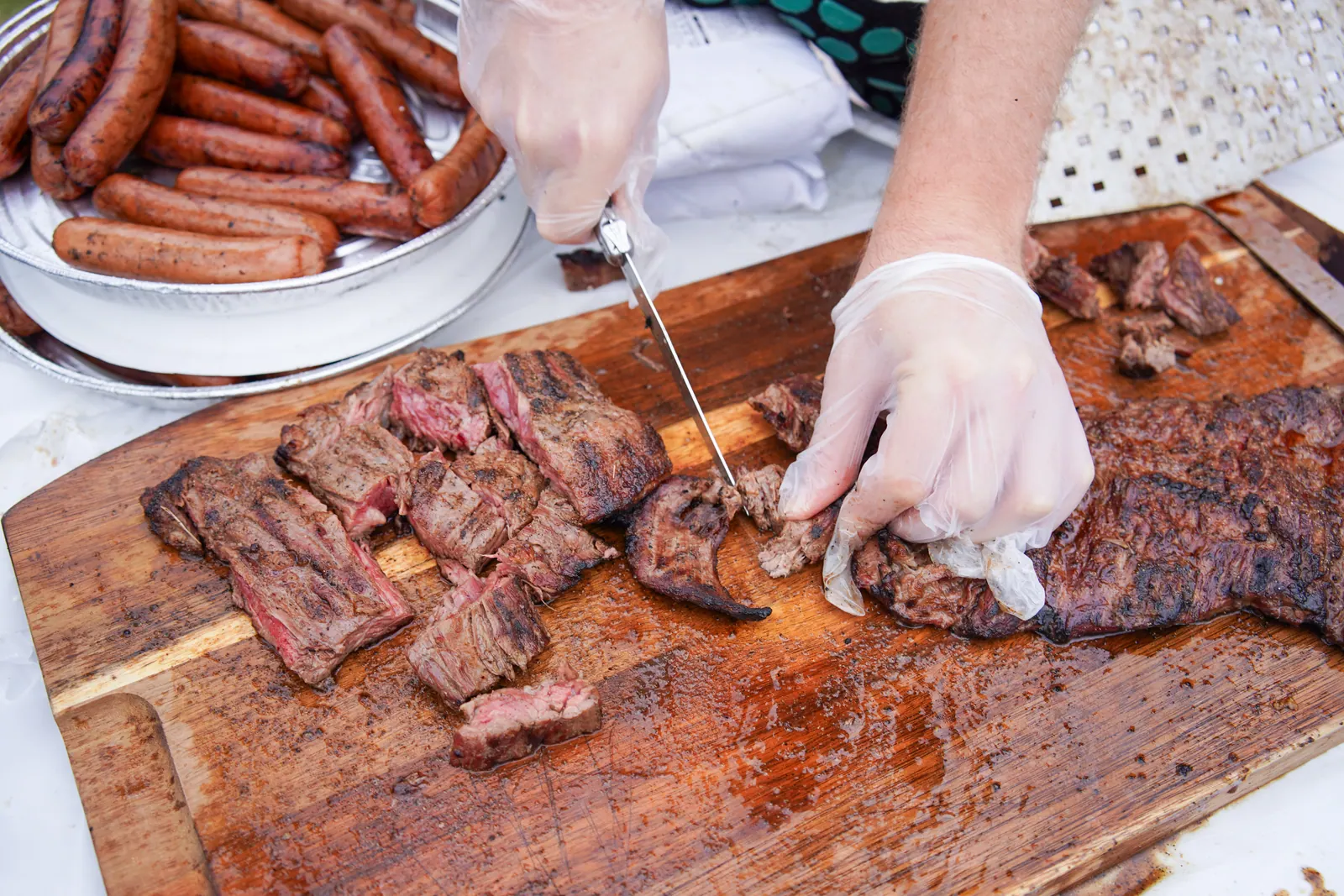 Gloved hands slice grilled steak on a cutting board beside a tray of sausages.