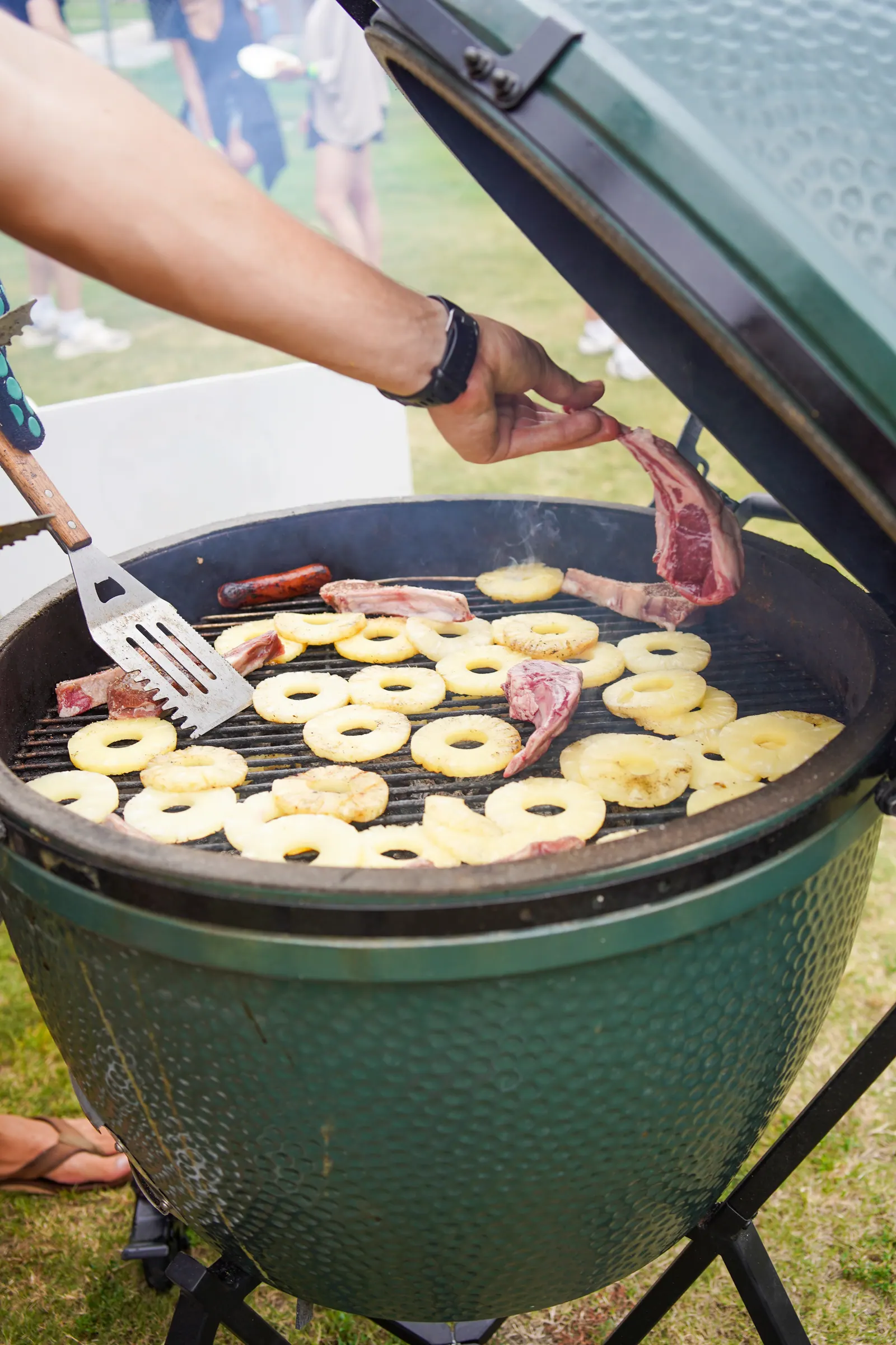 Pineapple rings and meat cooking on an open green ceramic grill.