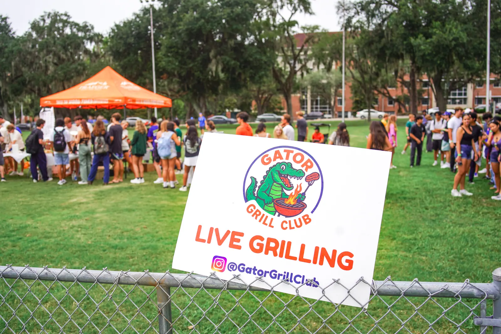A Gator Grill Club 'Live Grilling' sign on a fence in front of the tent and crowd.