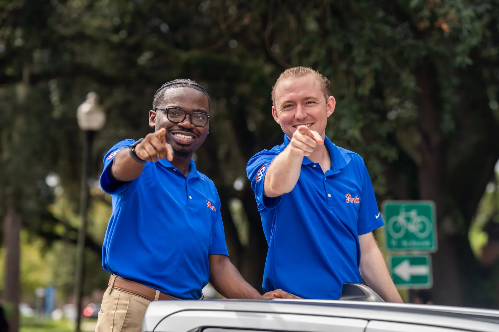 Two men in blue polos point at the camera from an open car roof.