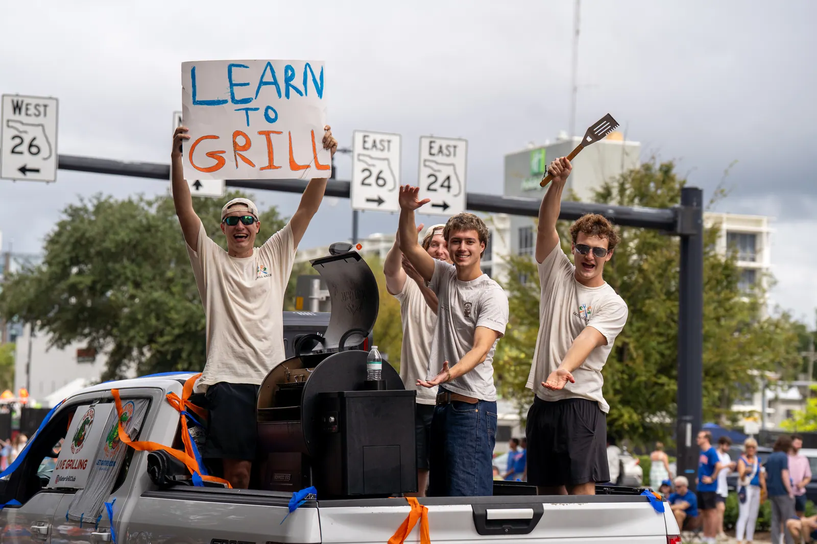 Students on a pickup truck hold up a 'Learn to Grill' sign and a spatula beside a smoker.