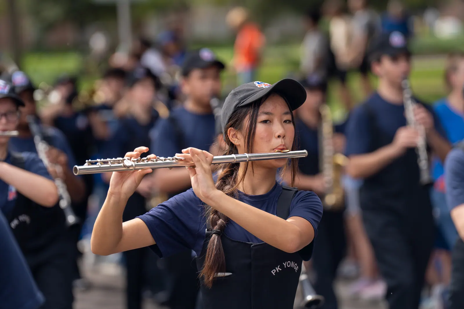 A marching band flutist in a cap plays as the band moves down the street.
