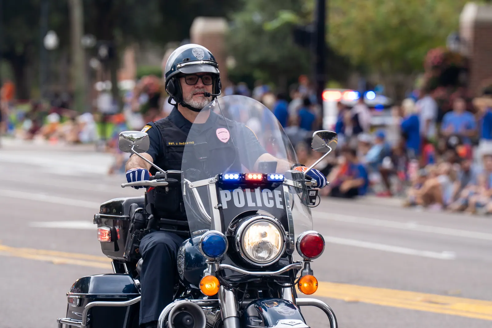 A police motorcycle officer leads the parade as crowds line the street.