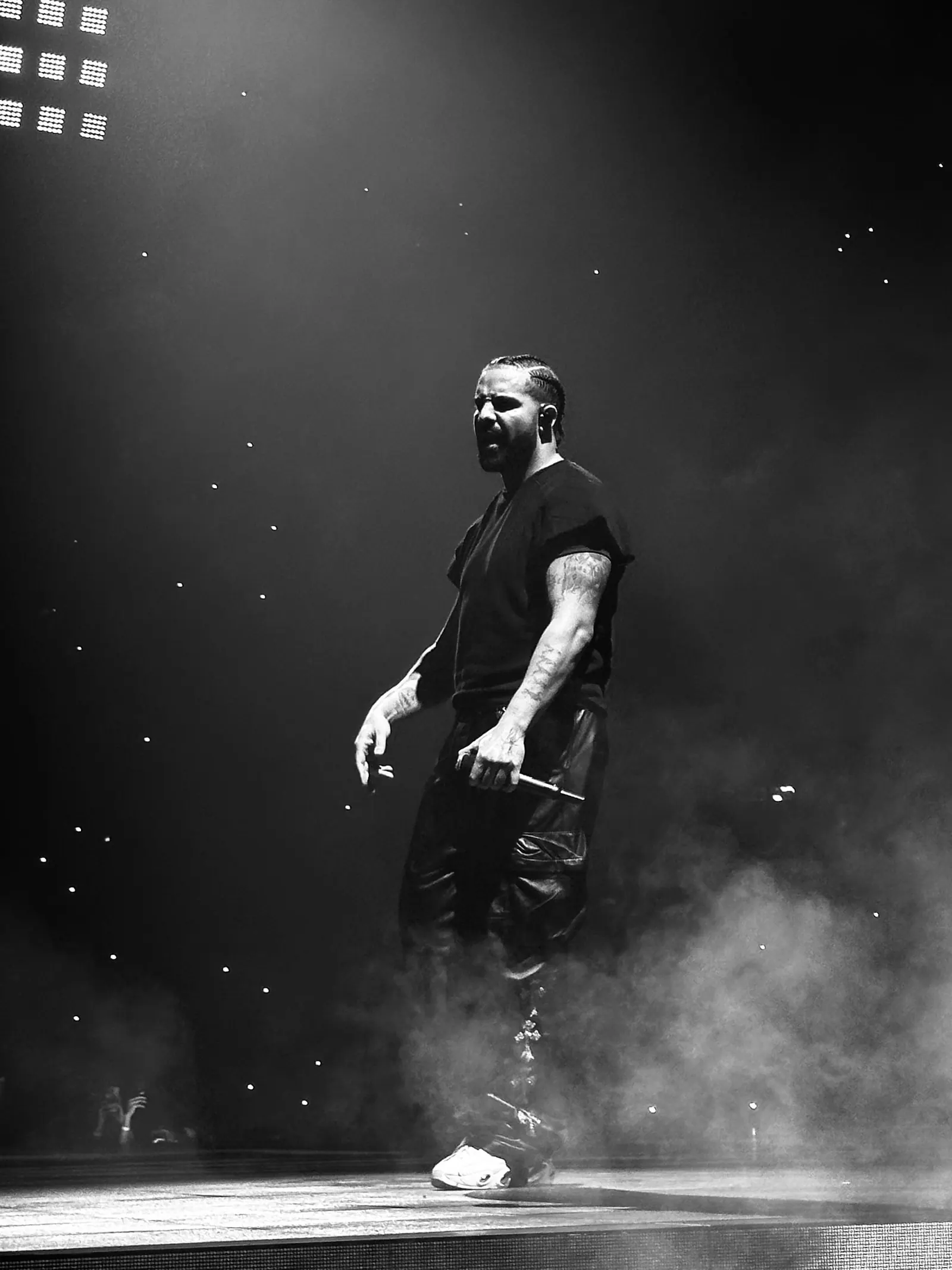 Black and white, a performer stands in stage smoke under arena lights, phone lights scattered like stars in the dark.