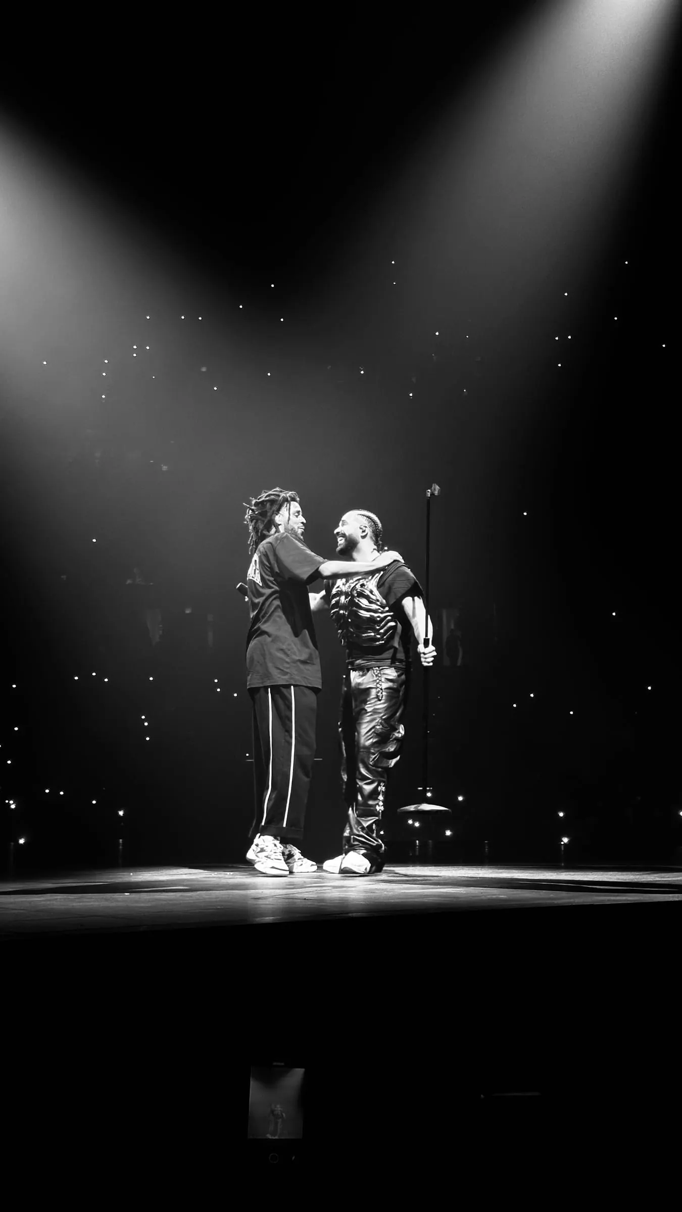 Black and white, two performers embrace in a single spotlight on an arena stage.