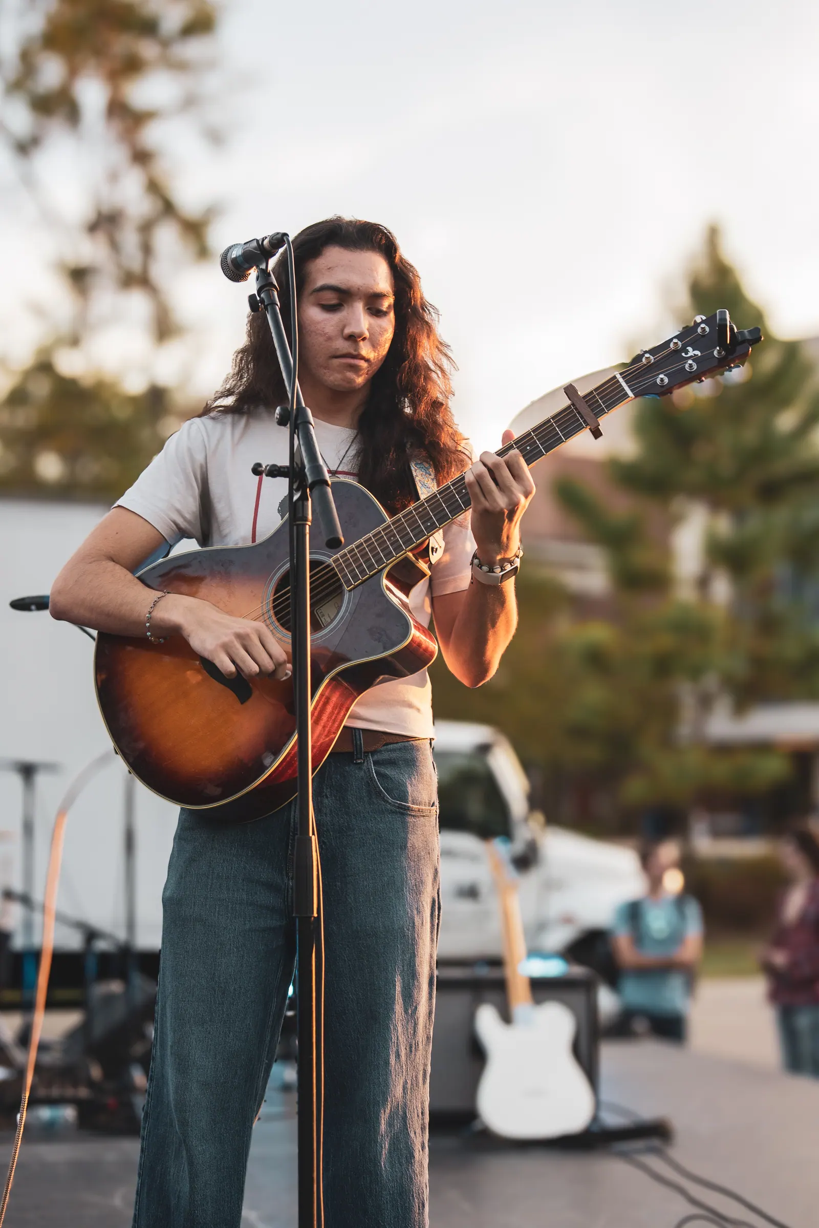 A long-haired guitarist playing an acoustic sunburst guitar at the microphone at golden hour.