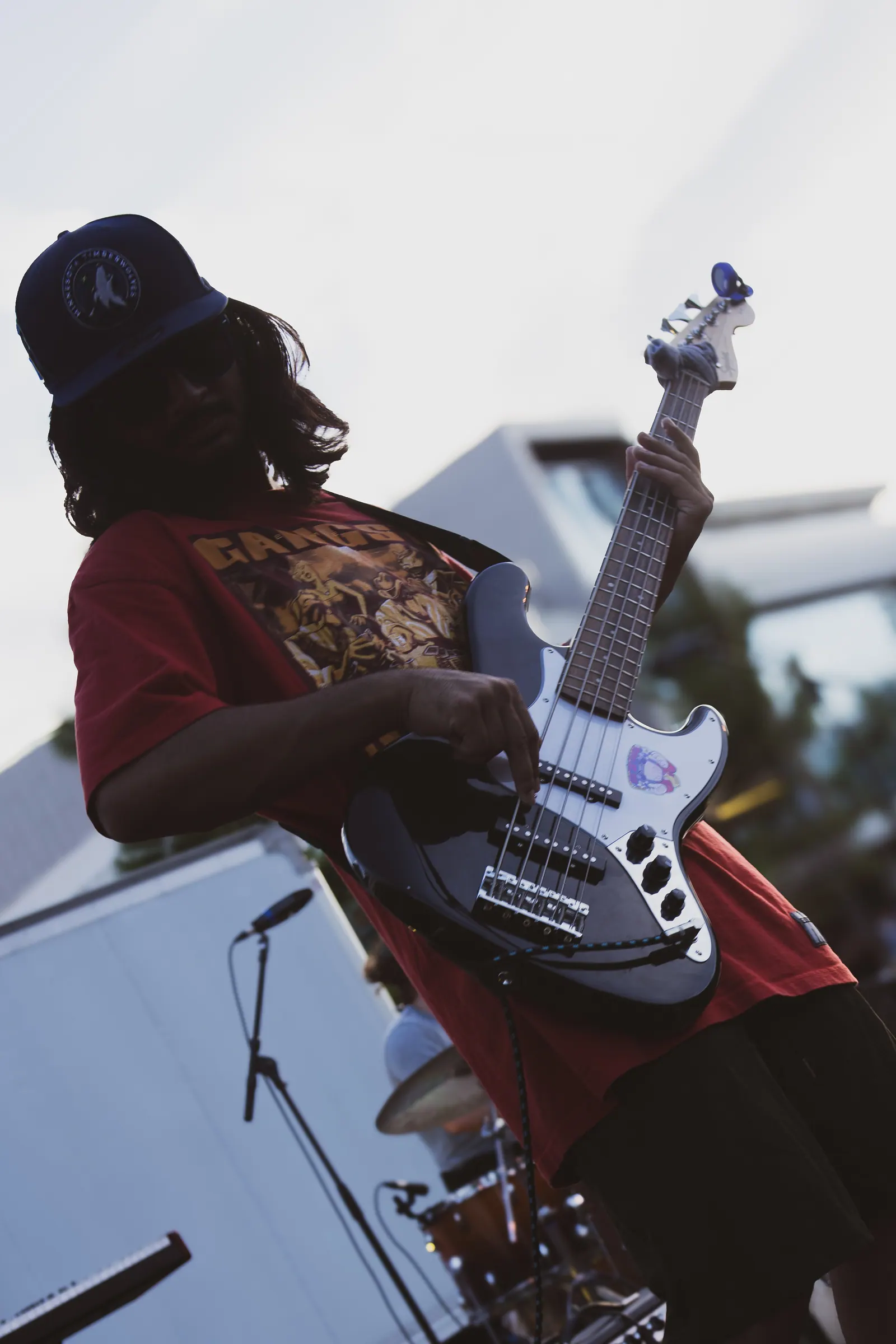 A bassist in a cap and red shirt, shot from below and tilted against a pale sky.