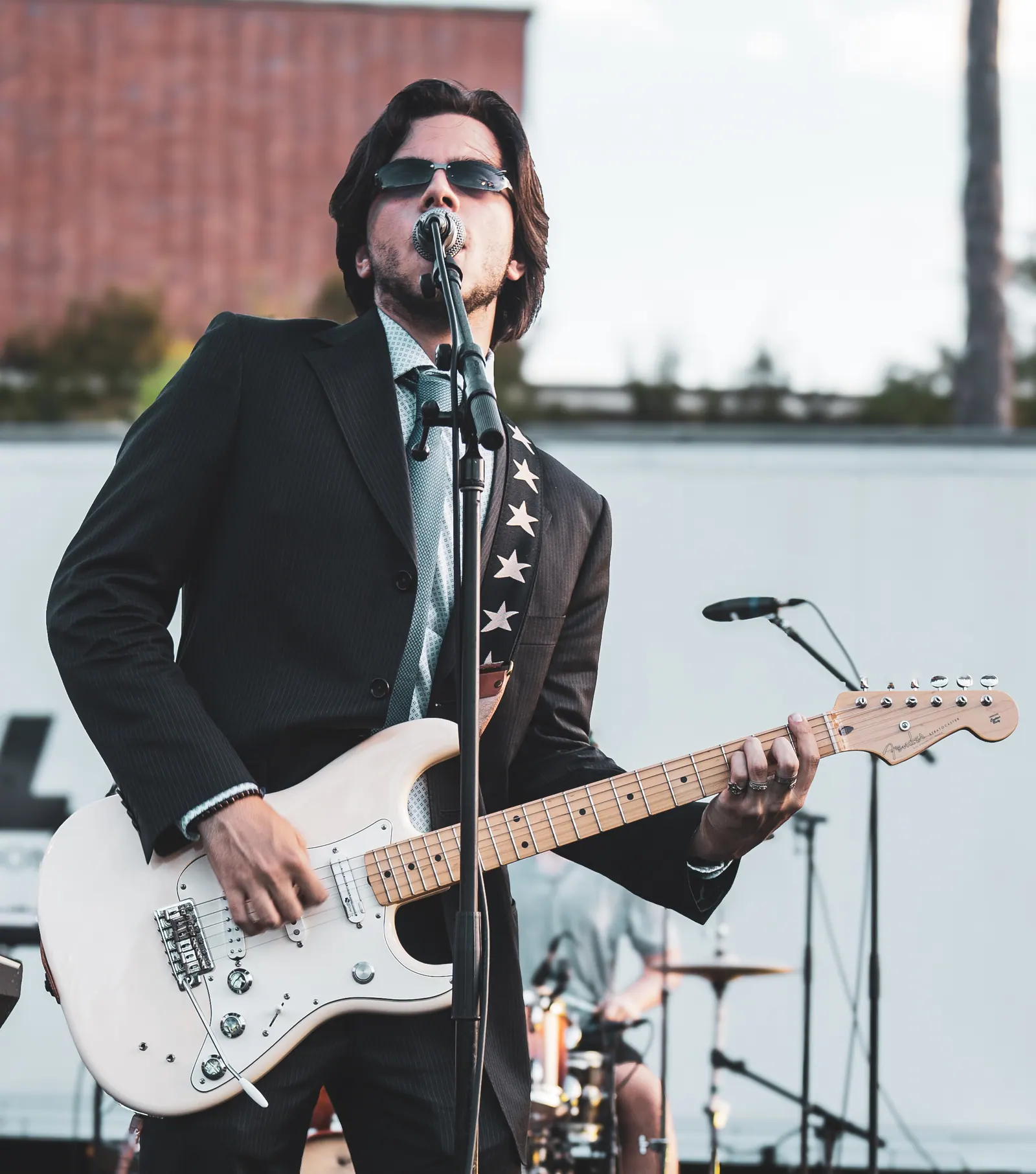 A singer in a black suit and sunglasses playing a white electric guitar at the microphone.
