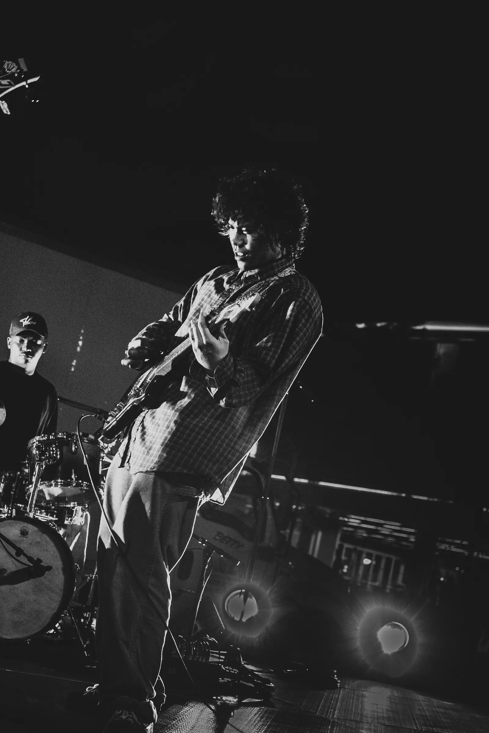Black and white, a guitarist leans into the guitar beside the drum kit on a dark stage.