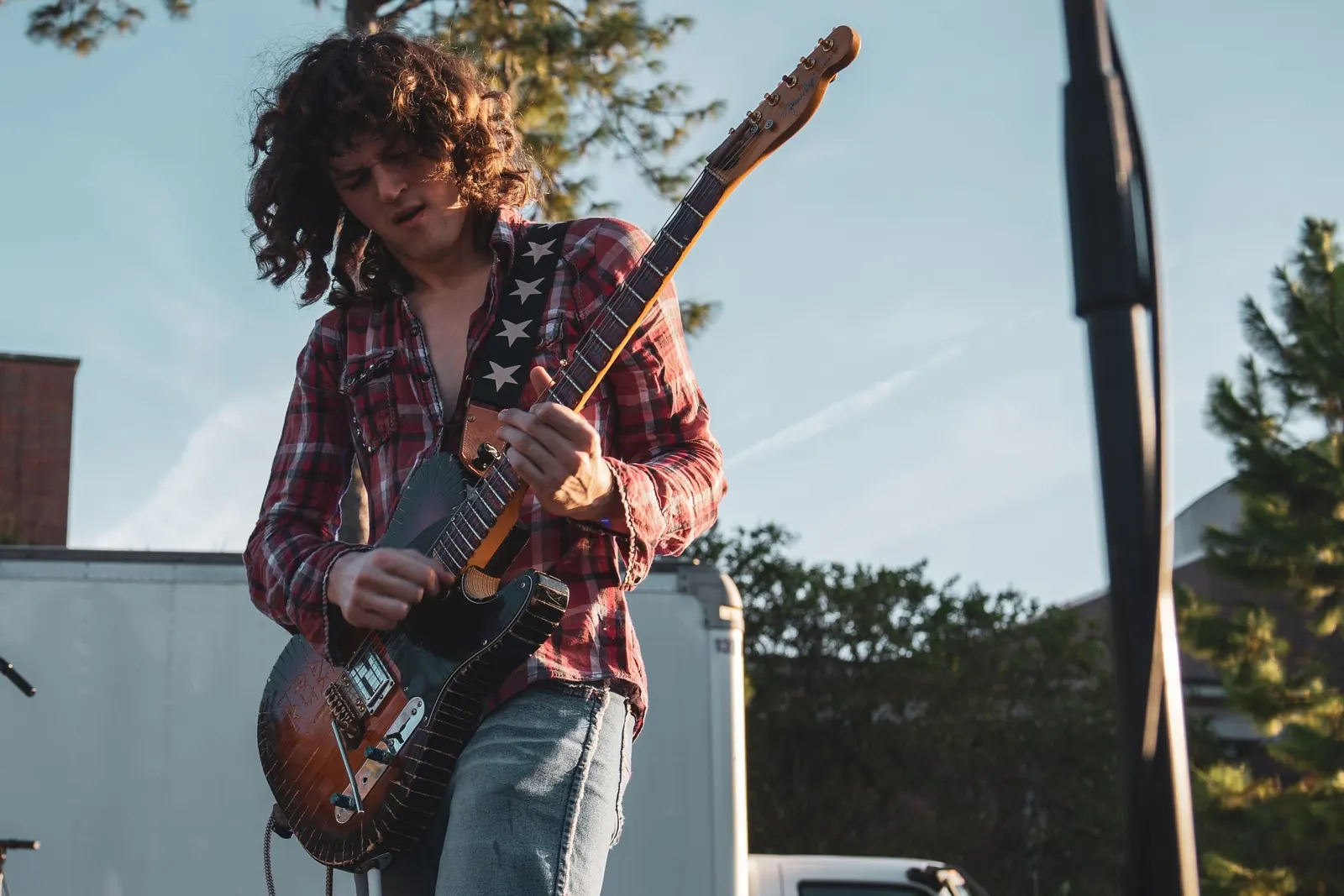 A curly-haired guitarist in a plaid shirt looking down at the guitar under an afternoon sky.