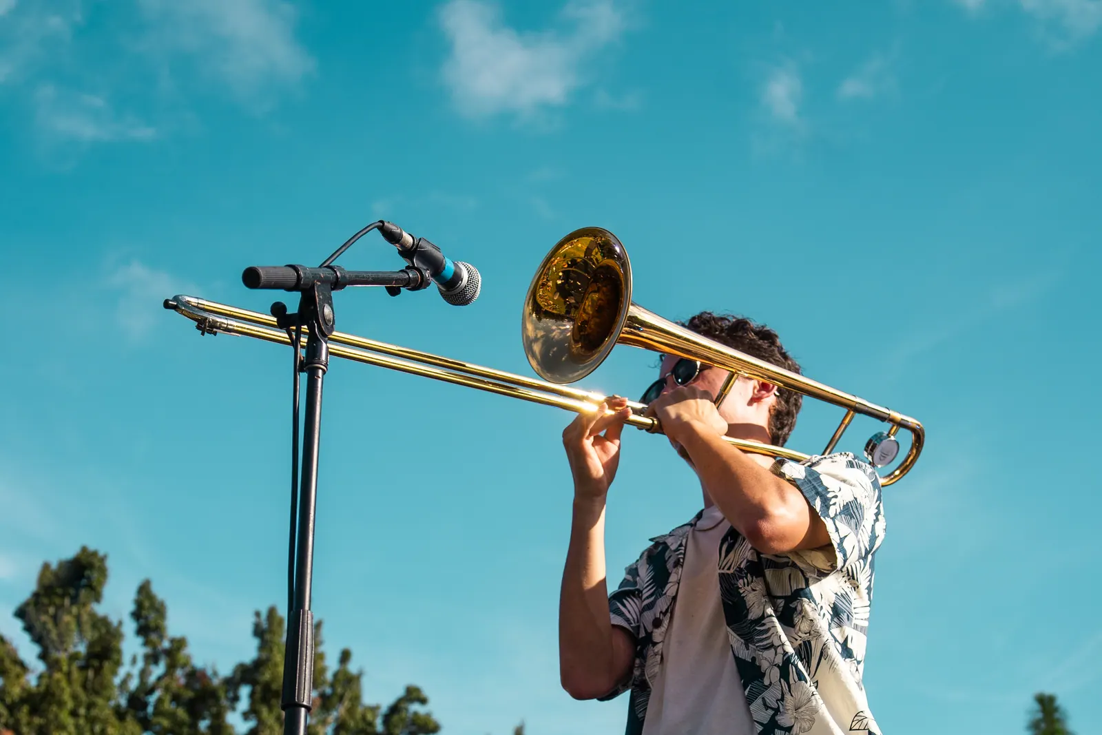 A trombonist in sunglasses and a floral shirt playing against a clear blue sky.