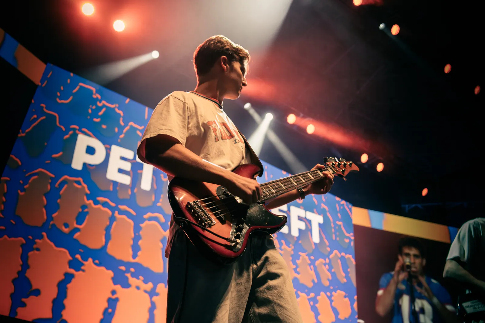 The bassist looks across the stage under warm spotlights, the band's name glowing on the screen behind.