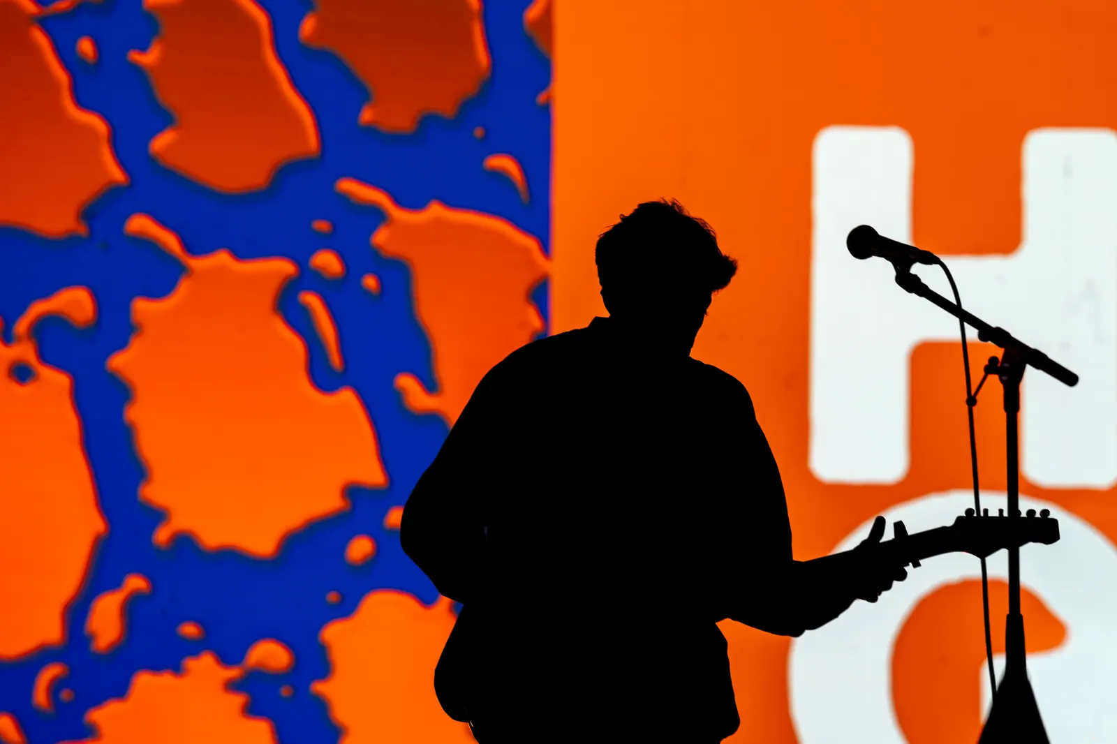 A guitarist in full silhouette against a bright orange and blue stage screen, microphone stand in the foreground.