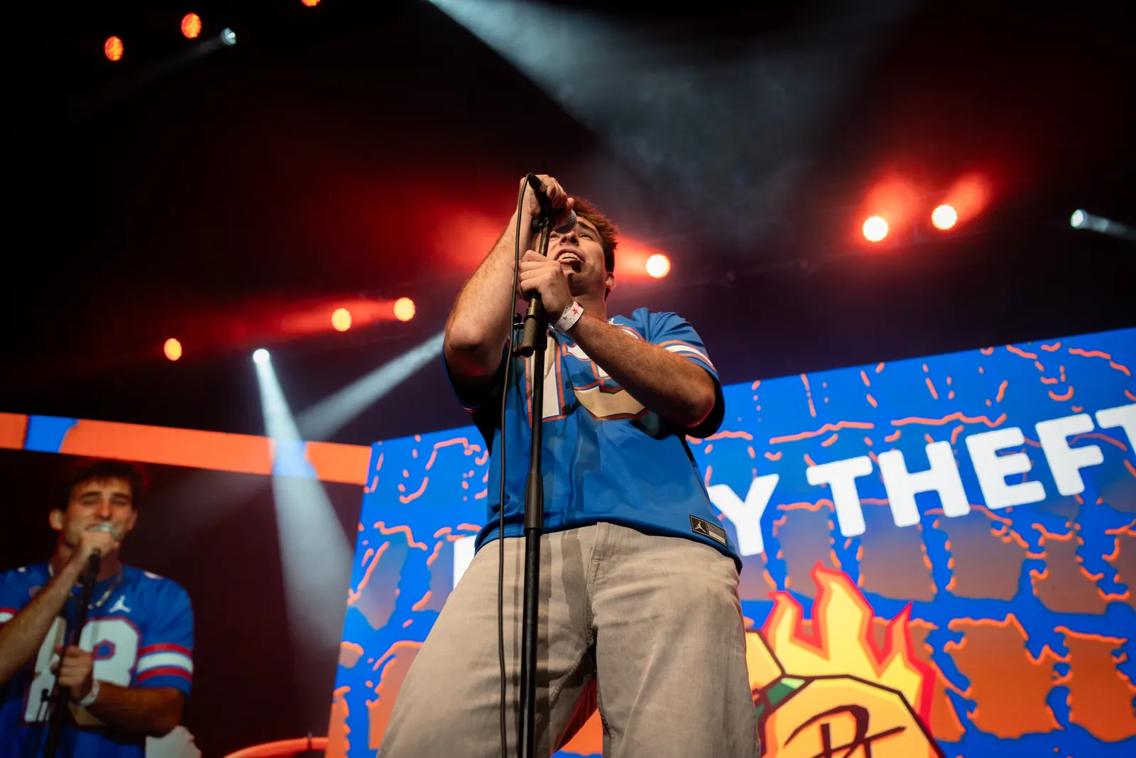 Low-angle view of the vocalist singing under red stage lights with the Petty Theft logo on the screen behind.