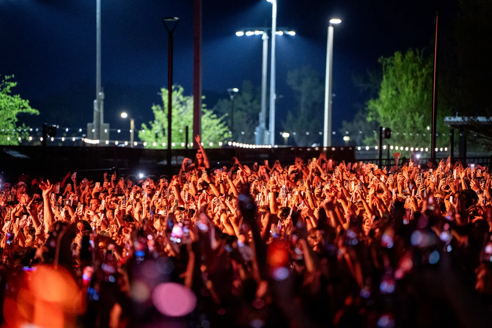A packed festival crowd lit red, hands and phones raised, under tall stadium lights.