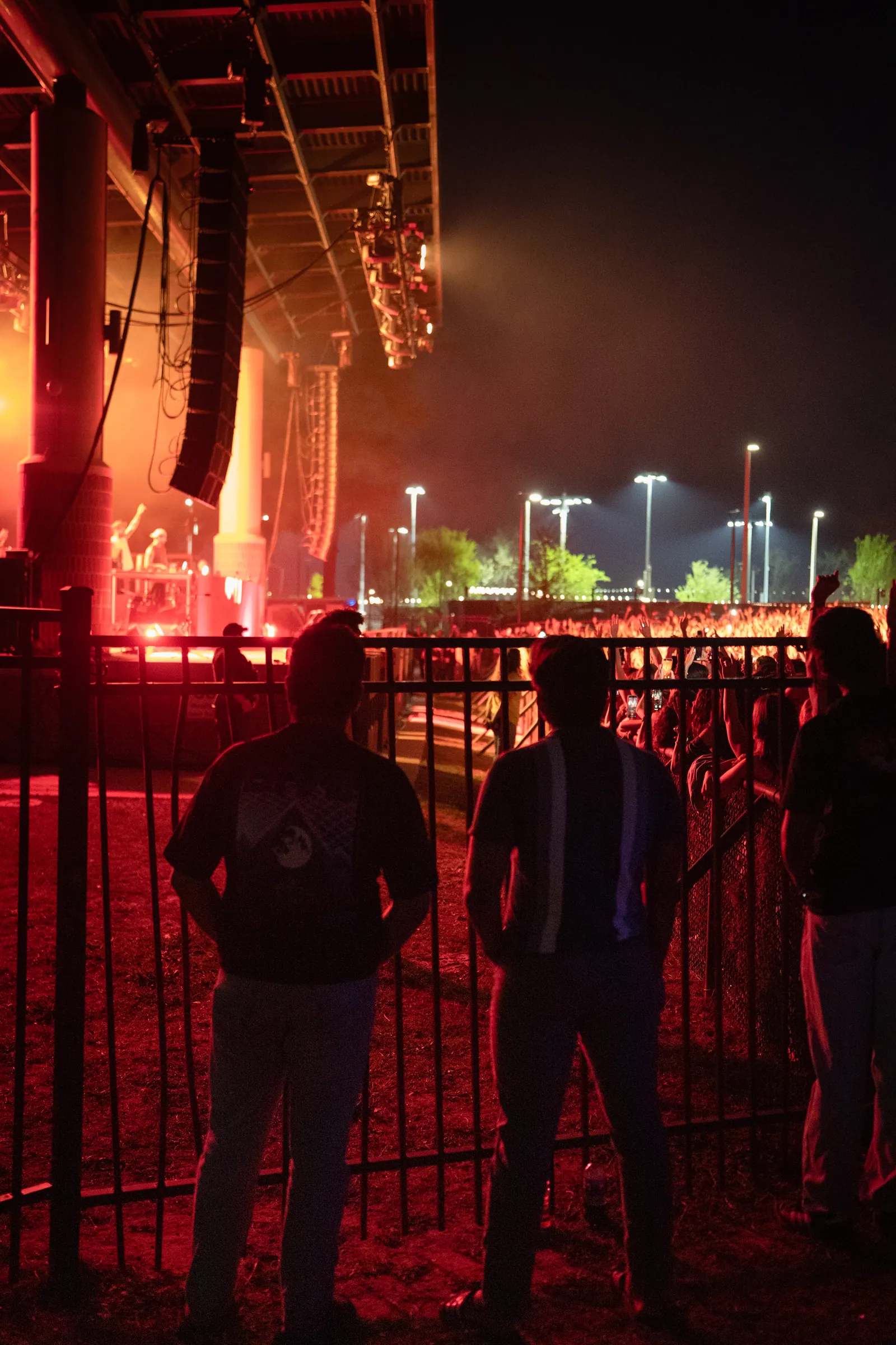 Three people in silhouette against a fence, the stage glowing red behind them.