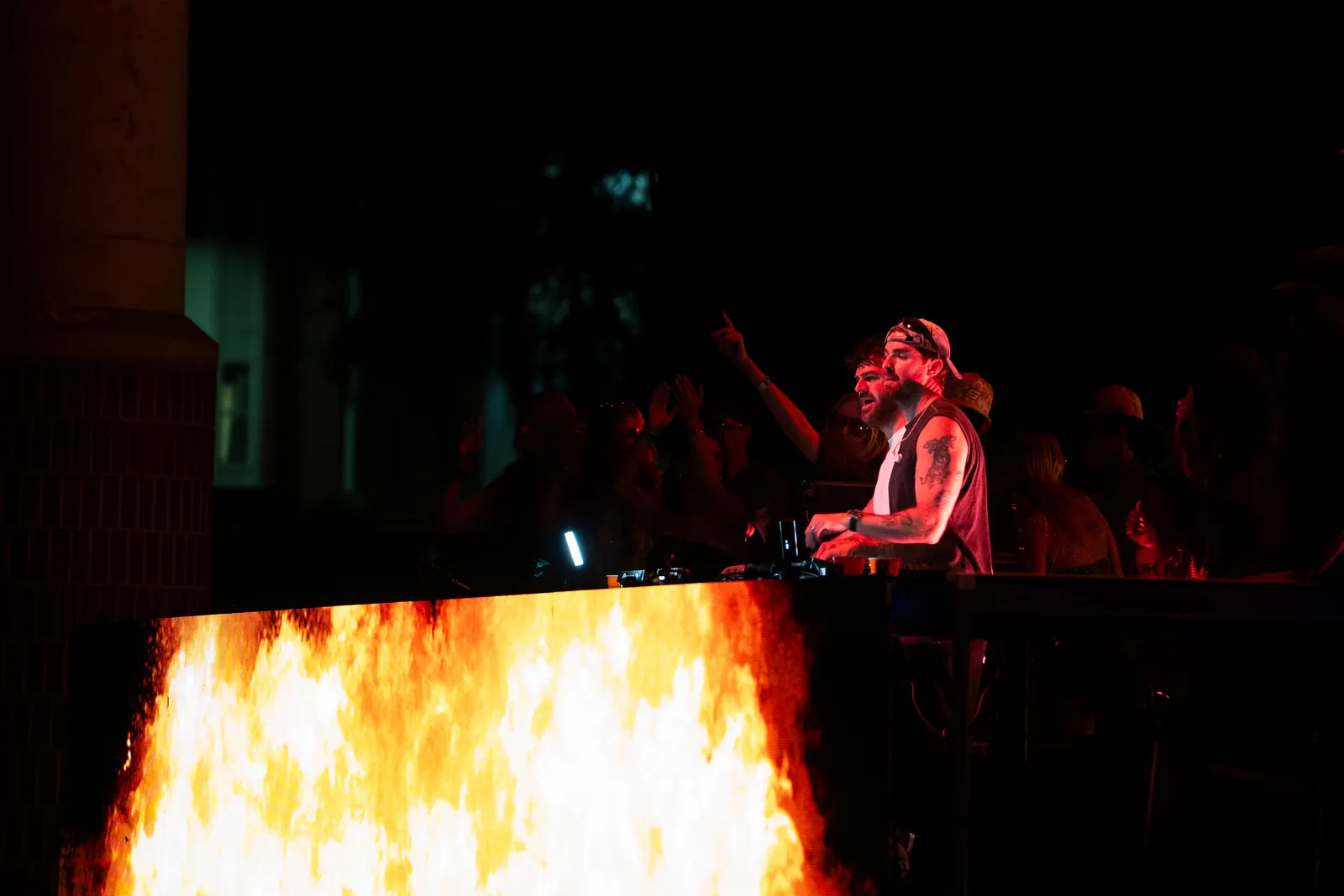 A DJ in red light points over the decks while a wall of fire plays on the screen below.