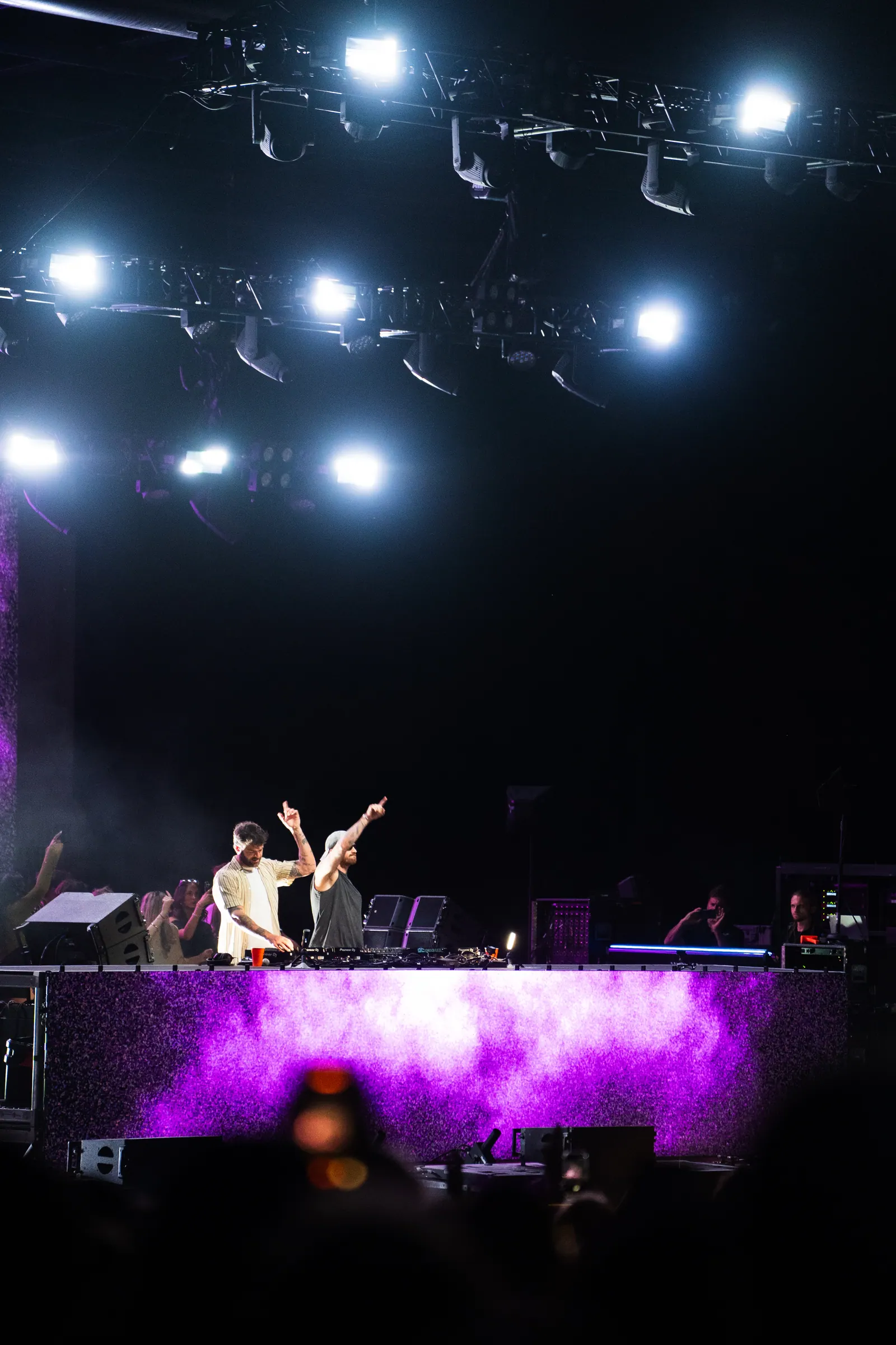 Two DJs on a distant purple-lit stage, one with both arms raised.