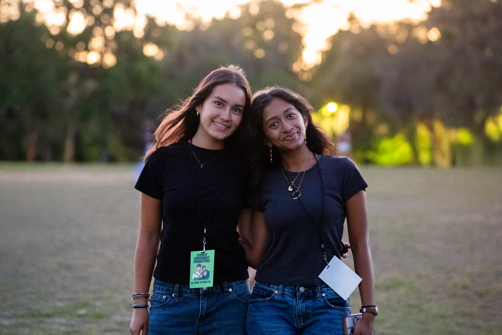 Two friends with festival wristbands pose arm in arm on the field at golden hour.