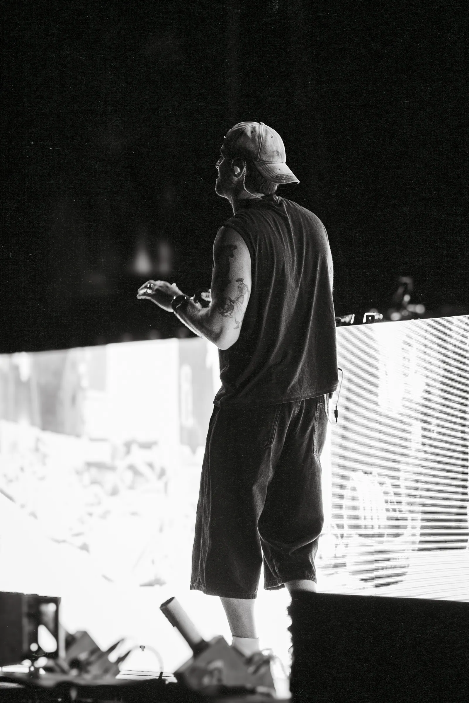Black and white, a performer in a cap stands on the stage edge in front of a bright screen.