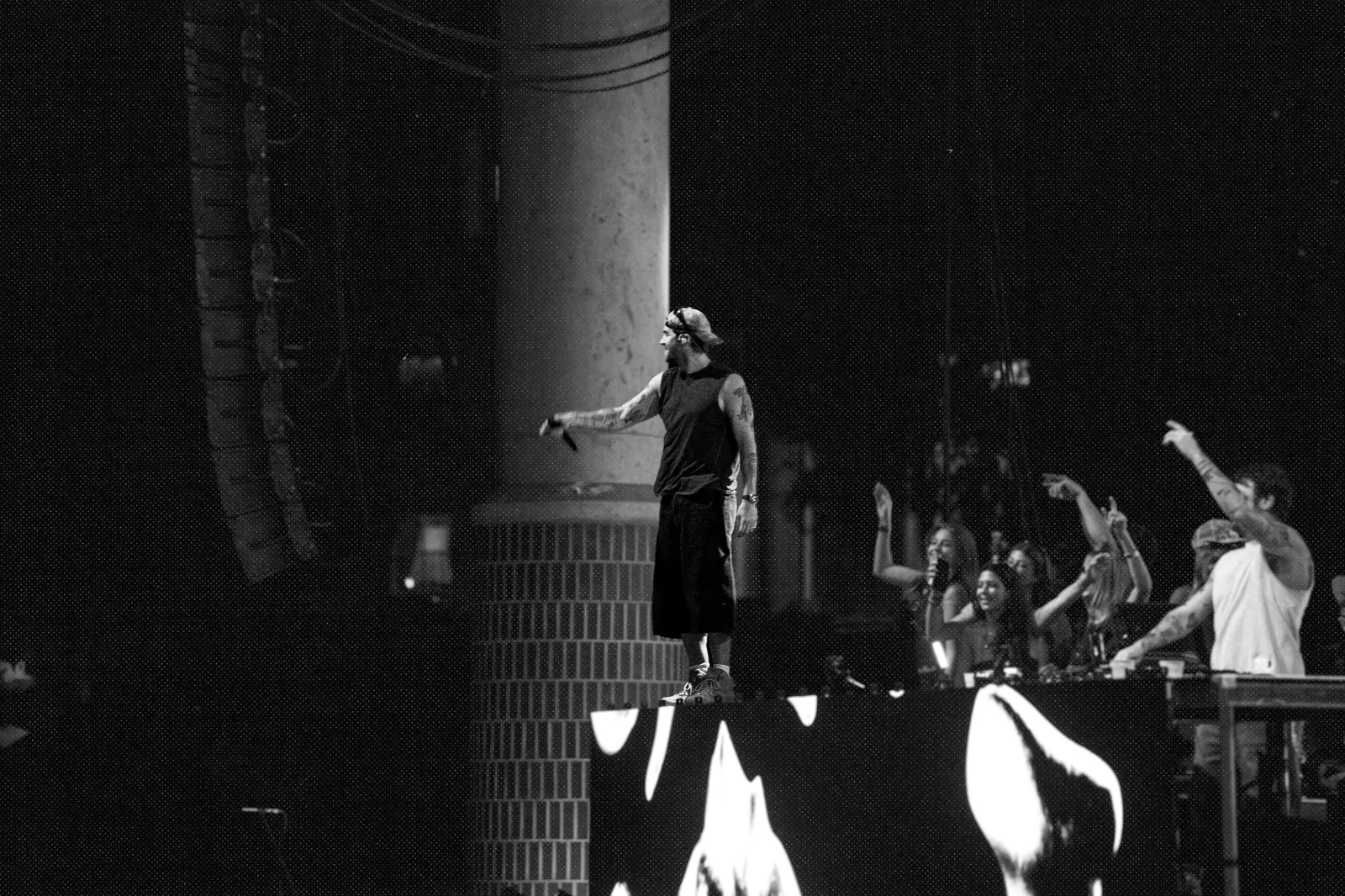 Black and white, a performer at the stage edge beside a brick pillar points toward the crowd.